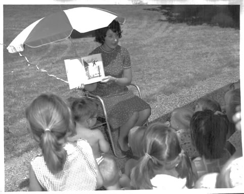 Black and white photo of a woman reading to children during Story hour on the front lawn.