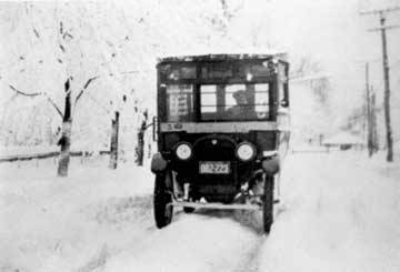 Black and white photograph showing an early bus making its way along a very snowy road.