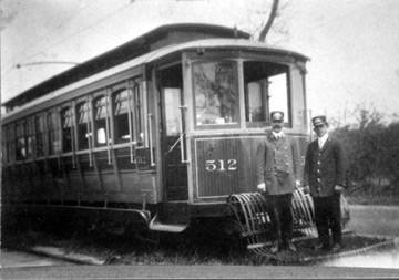 Black and white photograph showing a trolley car, complete with window shades, cow catcher, and electric wires overhead