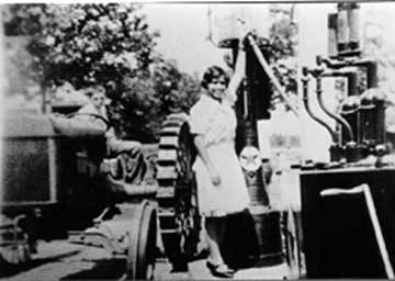 Black and white photograph showing a young woman in her dress gassing up the tractor at a very simple Texaco gas pump