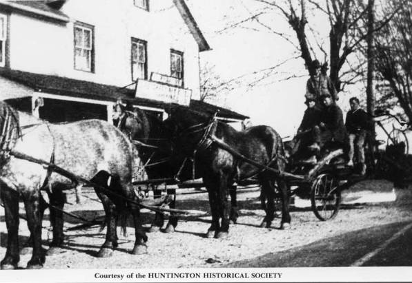 Black and white photograph showing a rig with four horses pulling four men aboard an open cart fitted with a scraping blade located just front of the rear axle