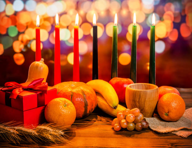 Picture of a candelabra with 7 lit candles, 3 red on the left, 3 green on the right and the center one is black. It represents the 7 life principles of Kwanzaa. The candelabra is placed on a table with various types of squash and grapes. 