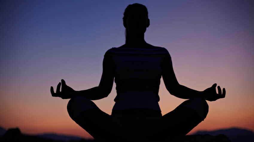 A silhouette of a woman meditating in front of a sunset.