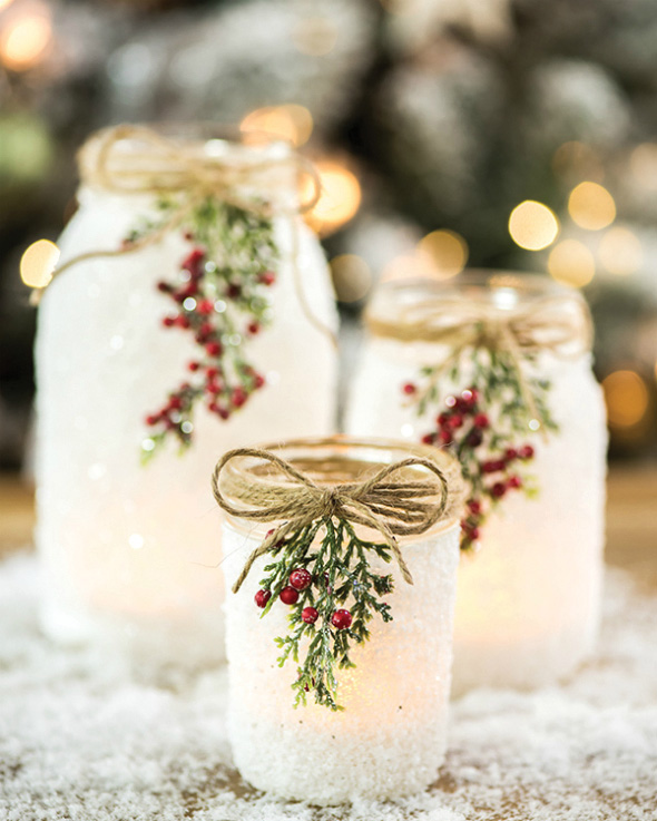 Image of a mason jar that looks like it has snow on it with a sprig of holly berries. It is lit up by a tea light candle inside. 