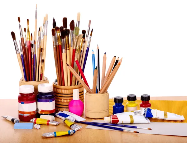 Photo of art supplies. A desktop with cups filled with paint brushes, colored pencils and on the desk, paper and paints.