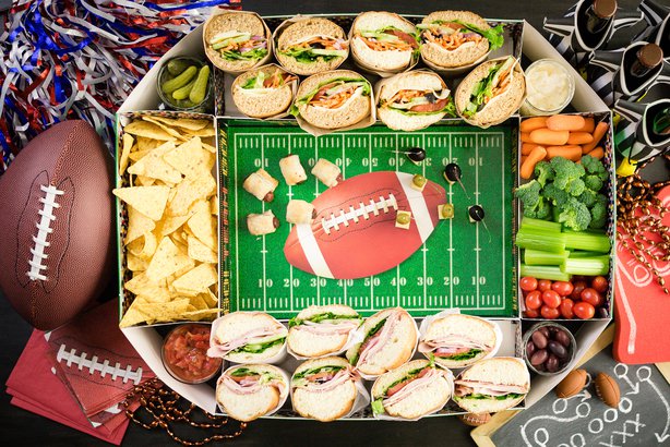 Image of a snack plate that looks like a football field. Around the tray are chips, veggie sticks, sandwiches and a real football off to the side. 
