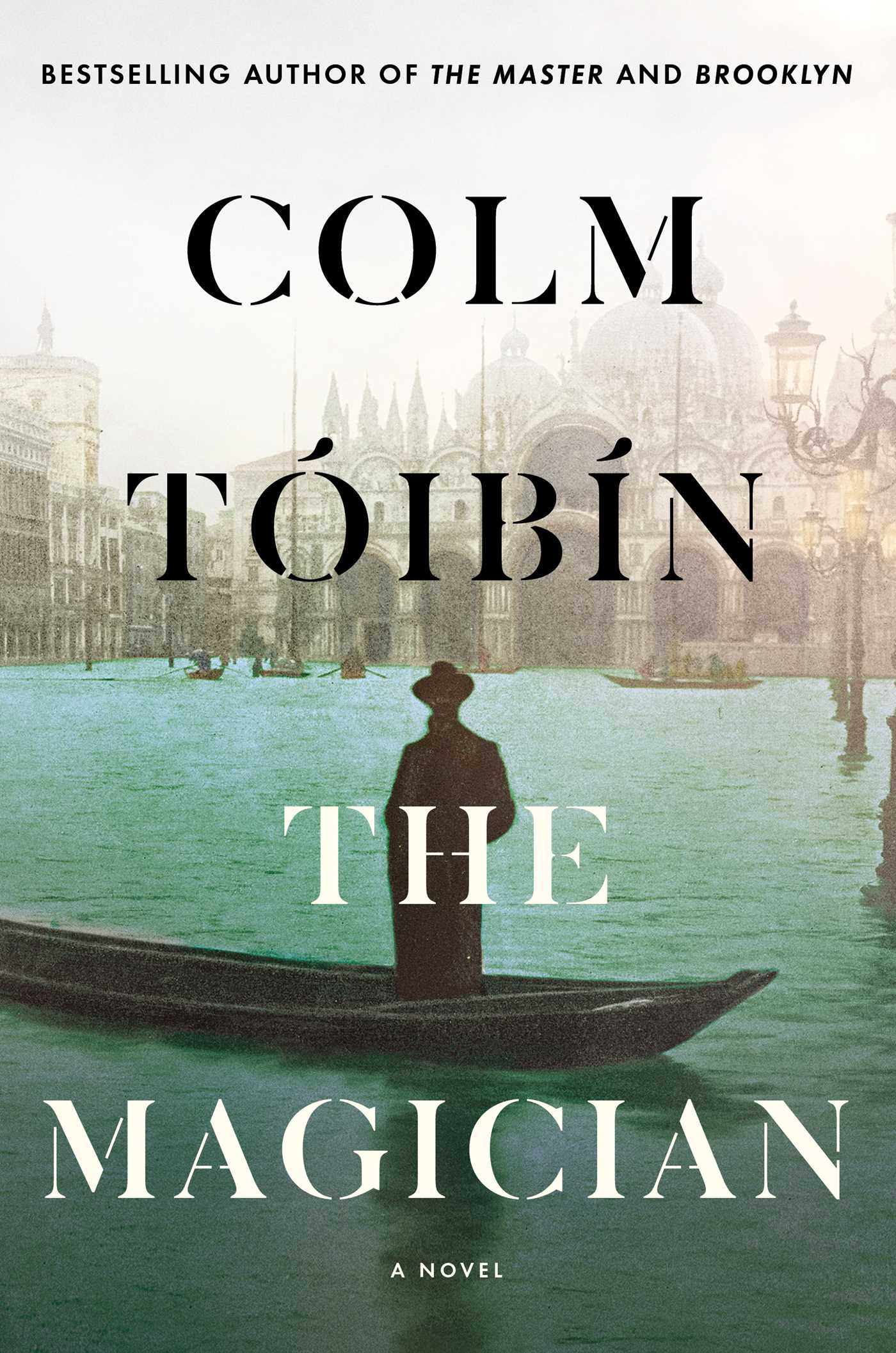 Image of the book cover of the novel The Magician by Colm Toibin featuring muted colors and a dark figure of a man in a hat and long coat standing in a gondola surrounded by architecturally beautiful buildings on what appears to be the waterways in Venice, Italy. Beautiful ar