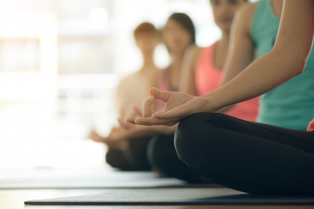 Image of 4 people sitting on yoga mats one behind the other in a sitting yoga pose. The image is clear in the front of the photo and blurred as it goes back to conceal the faces of the people. 