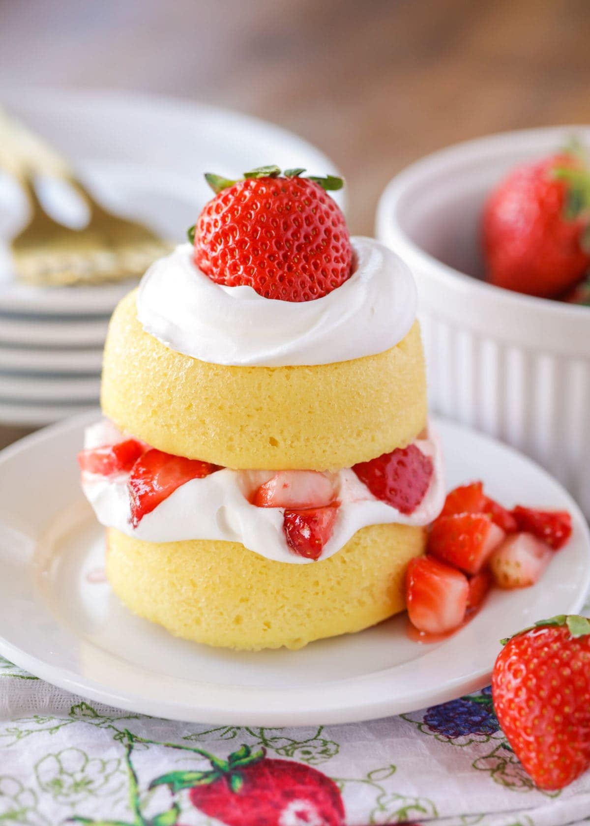 Image of a small strawberry shortcake. Two small round vanilla cake portions stacked with whipped cream and strawberries in between and on the top, more whipped cream and a whole strawberry. Some cut up strawberries on the side of the plate.