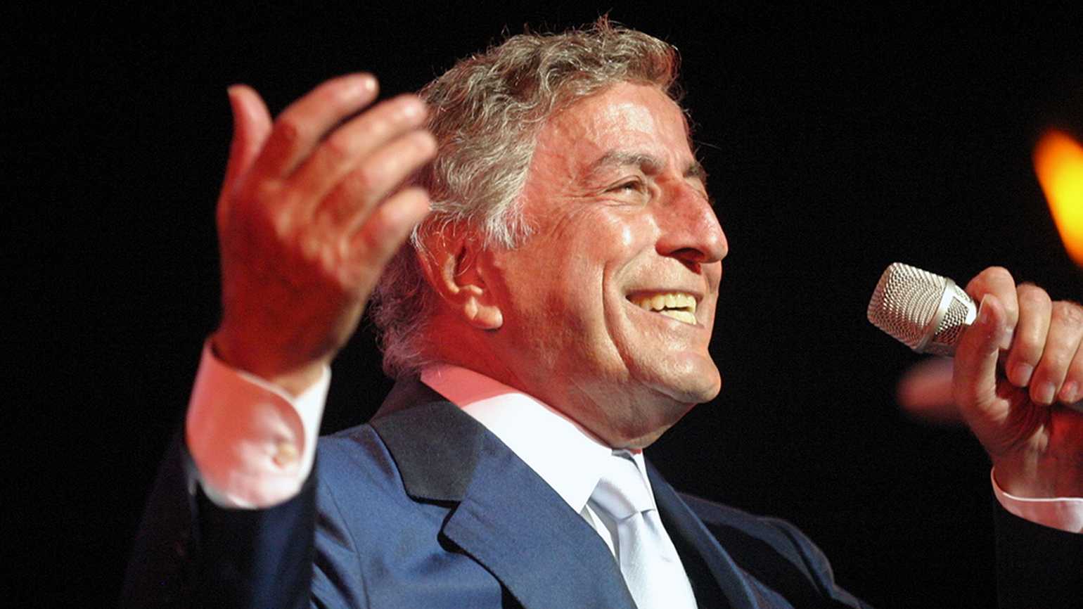 Image of Tony Bennett in his senior years performing. His left hand holding the microphone, his right hand uplifted and a smile on his face. 