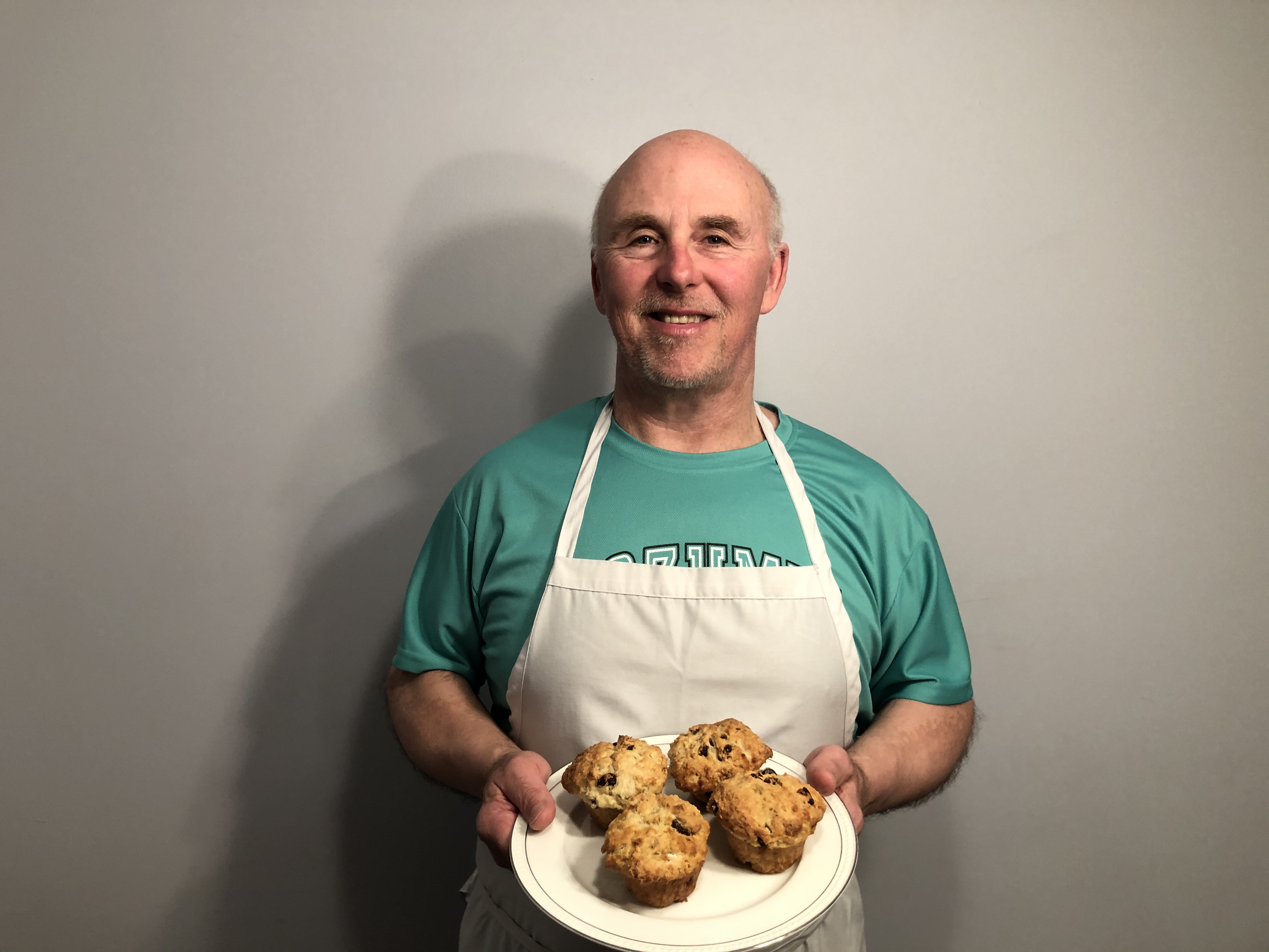 Man dressed in a white apron holding a plate of muffins. 