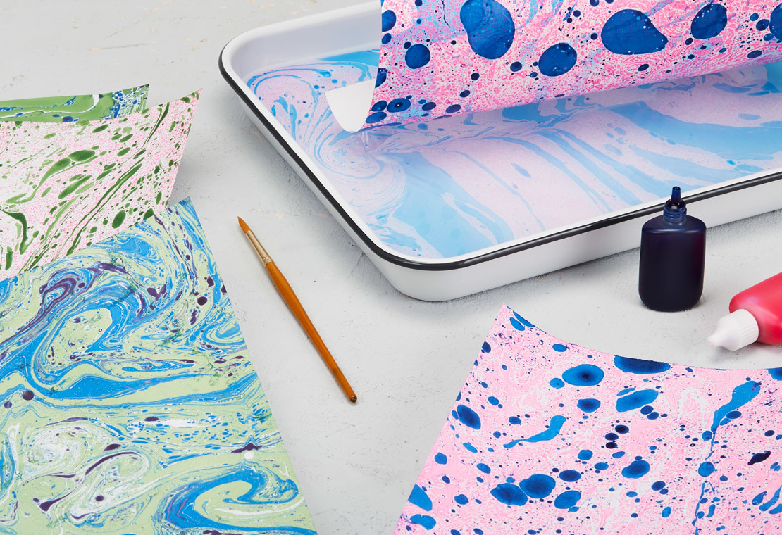 Image of a tray with food coloring in it. Pieces of paper being dipped face down in the tray for the paper to absorb the color to create a marbling effect.