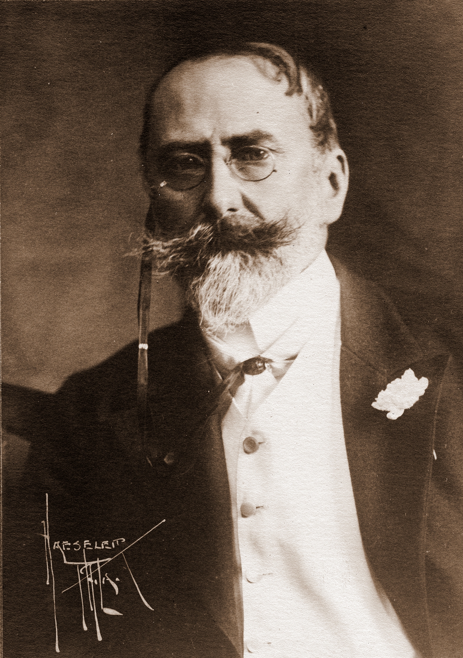 Old photograph of William Merritt Chase. Man in formal wear with spectacles. Has an untrimmed moustache and a white beard.