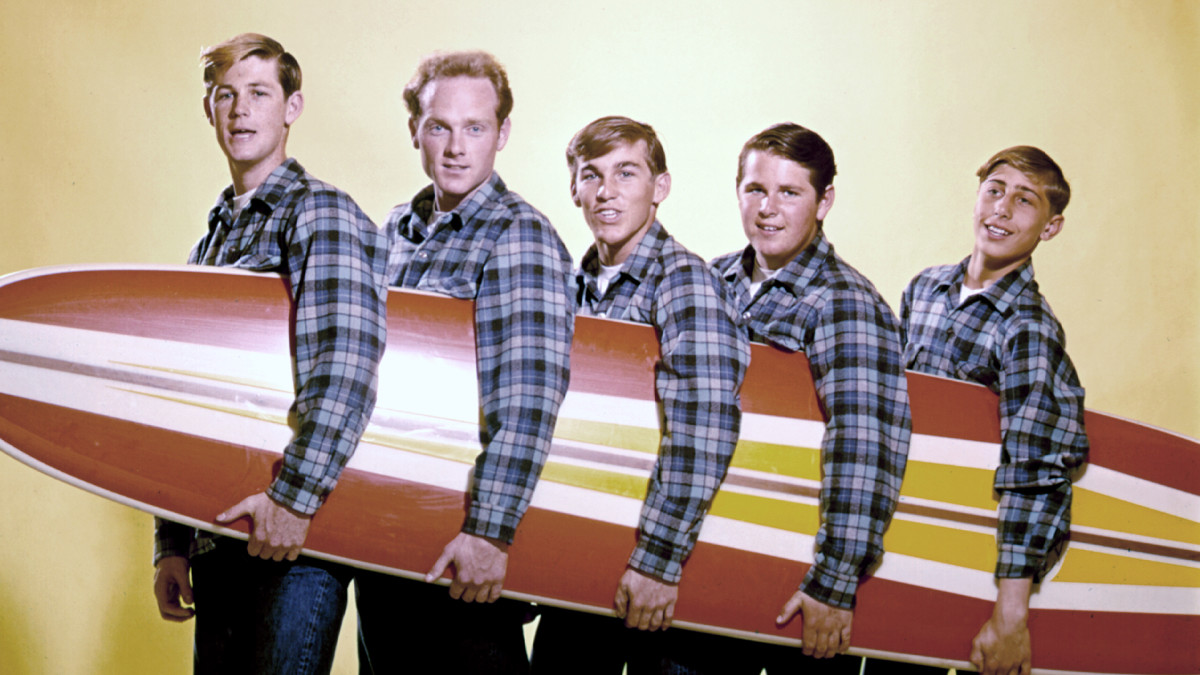 Image of the Beach Boys group. It is a picture when they were young. They are wearing matching plaid long sleeved shirts and are holding a surfboard. 