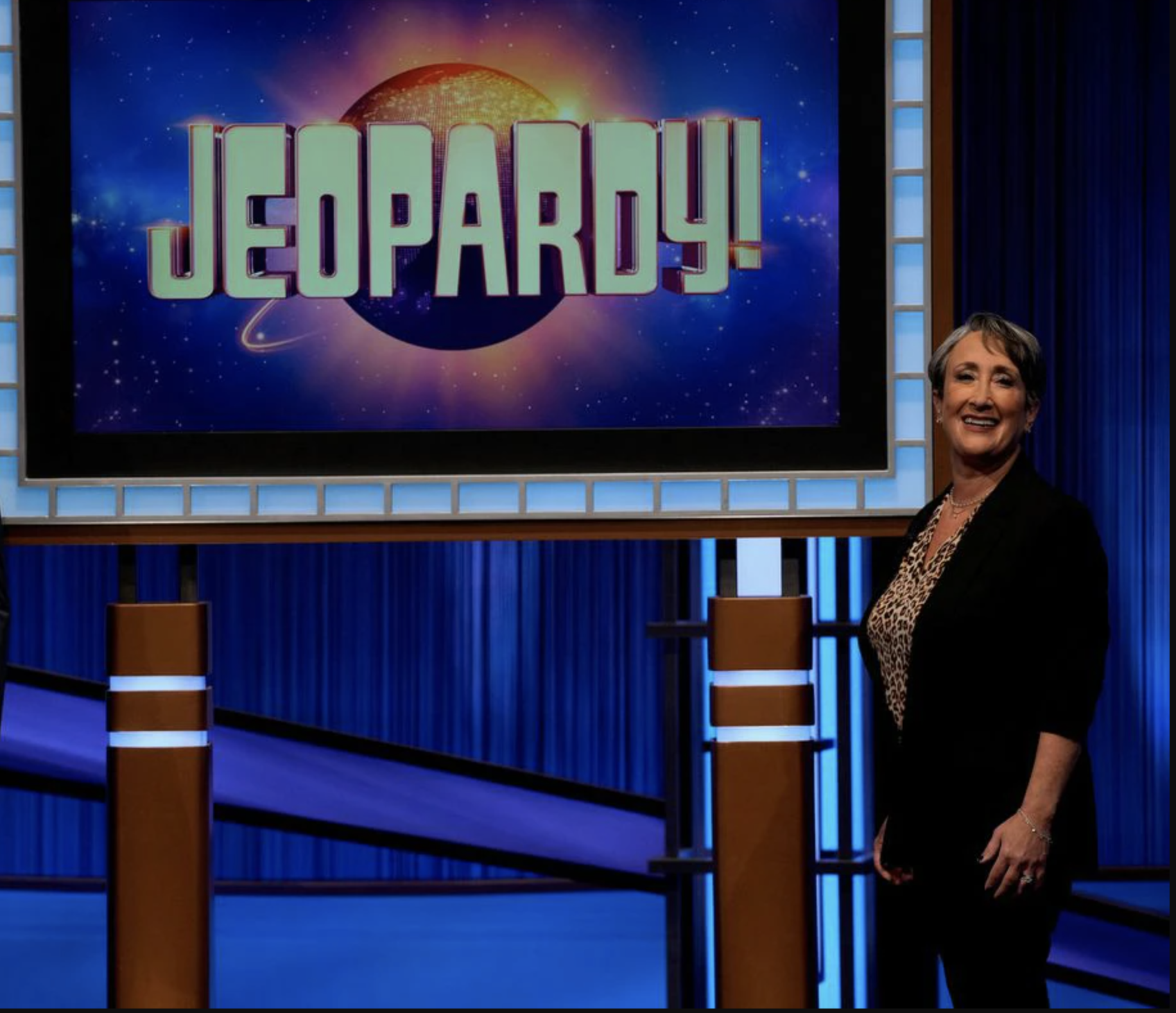 Image of a woman standing next to a screen with the Jeopardy Logo on it. 