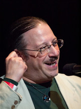 Photo of the presenter, Tony Traguardo featuring a male with a mustache sitting in profile, wearing eye glasses, a tan jacket. His right hand on his right ear and he is smiling. 