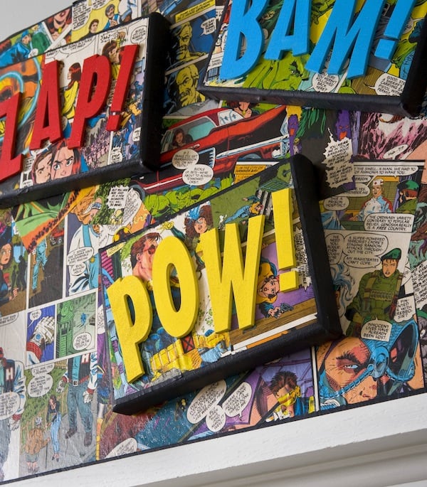 Image of a wall with comics pasted on the wall. Three picture frames that have comics and each with a different super hero word like Zap, Bam and Pow!