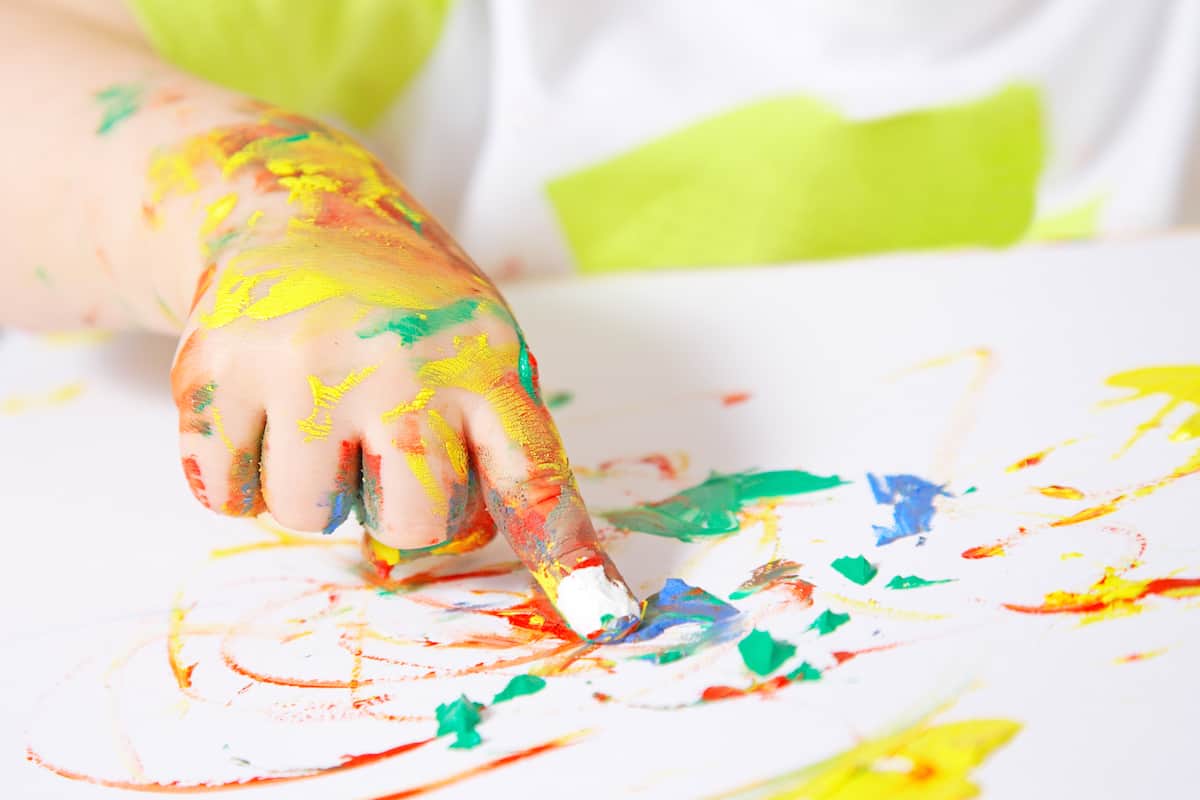 image of a small child's hand painting with the fingers. Colorful paint is on the paper and on the child's hand.