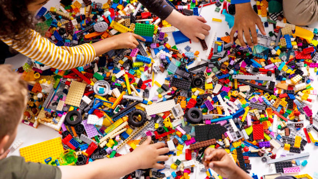 Image of many multicolored LEGOS and children's arms and hands reaching for the pieces. 