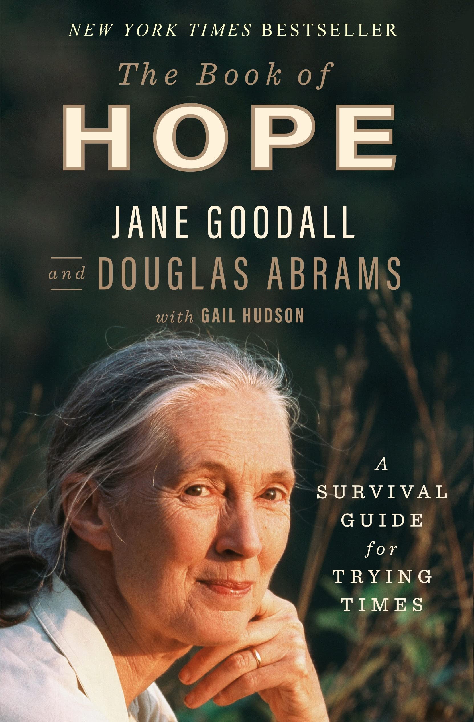 Book cover for the book Book Title: A Book Of Hope: A Survival Guide for Trying Times by Jane Goodall and Douglas Abrams with Gail Hudson featuring a photo of Jane Goodall. A woman of a mature age with grey hair pulled back in a ponytail. Her left hand is by her jawbone. She has a gold wedding band on her ring finger. 