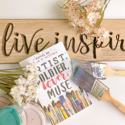 image of a novel titled "Artist Soldier lover Muse" laying atop a wood board that reads "Live inspire" with white flowers and paintbrushes on top of the book.