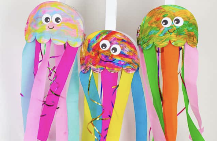 Image of 3 jellyfish windsocks hanging from white bands, made from pinks, blues, yellows, and greens with large cutout eyes.