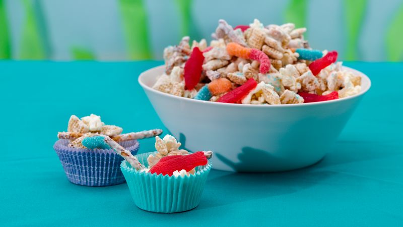 Image of trail mix, gummy worms, and Swedish fish in a bowl and cupcake cup holders on a blue table. 