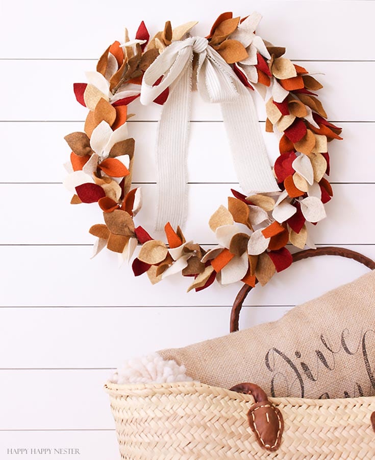 A felt fall themed wreath hung on a white brick wall in front of a chair with a pillow on it.