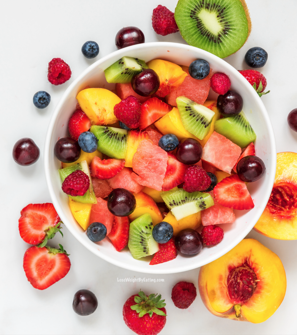Photo of all different kinds of colorful fruit in a white bowl. Some of the cut up pieces of fruit are surrounding the bowl on the table.