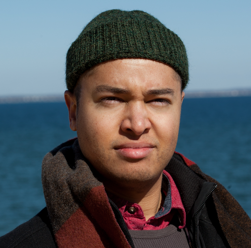 Headshot of a man, Jeremy Dennis, in a green beanie looking past the camera with a background of the ocean behind him. He is wearing wintery clothing and a thicker coat.