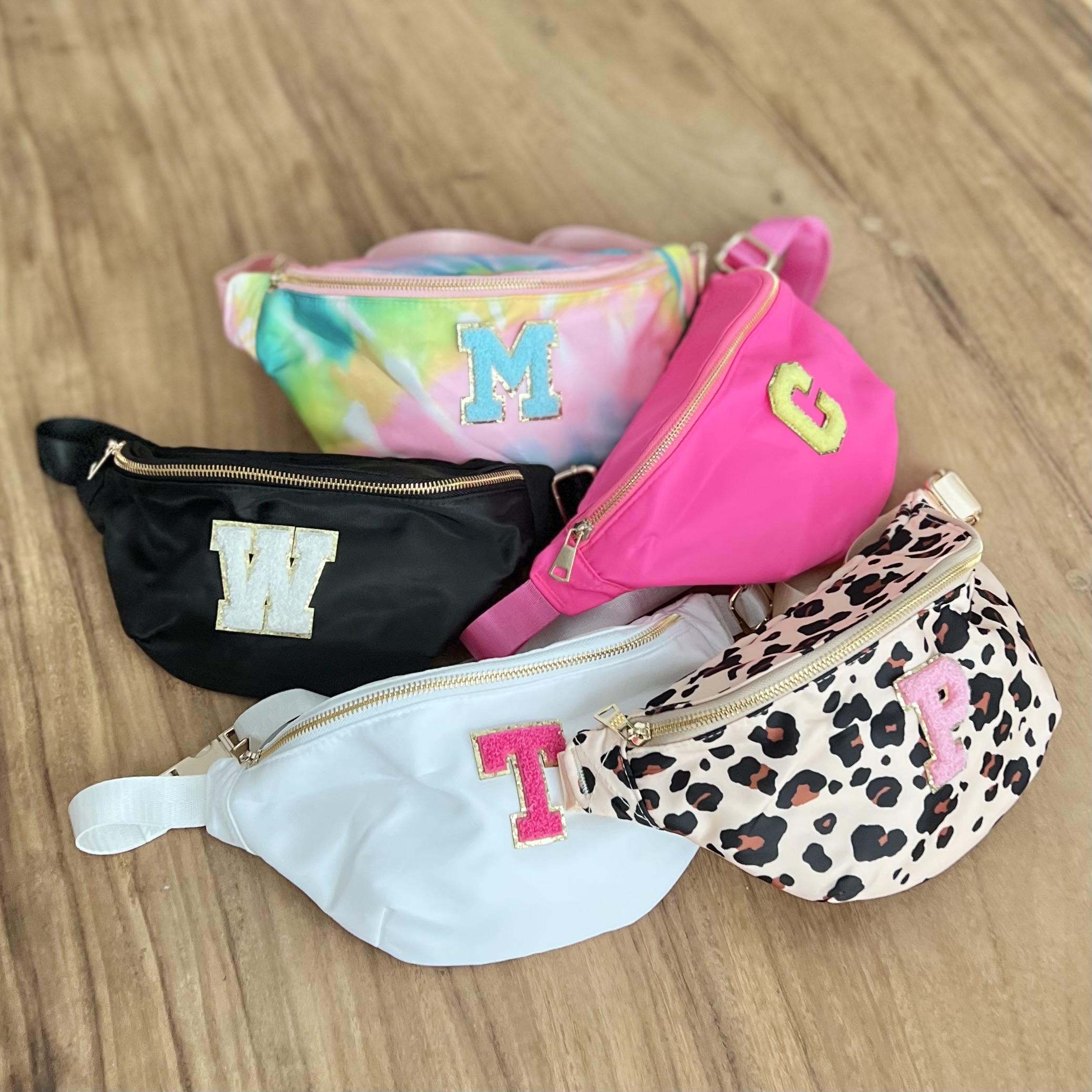 Image of 5 fanny packs on a wooden floor, each a different color with various patch letters on each.