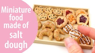 image of miniature sculptures of croissants, cookies, and danishes, as well as someone's hand holding a miniature sculpted cinnamon roll between their thumb and forefinger. A pink semi circle on the left reads Miniature food made of salt dough.
