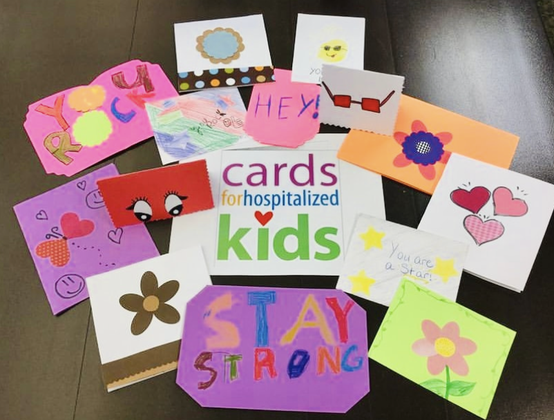 Cards for Hospitalized Kids logo on a piece of paper, laying flat amidst 15 handmade cards, drawn in crayon and marker, with phrases such as YOU ROCK and STAY STRONG written on them in multiple colors.