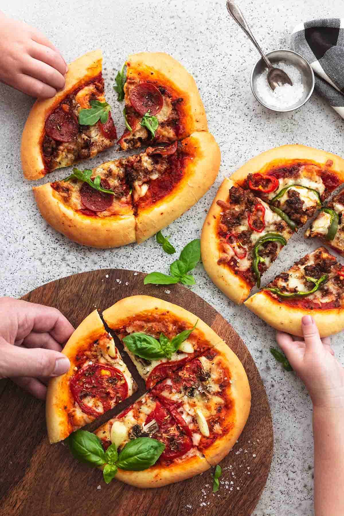 Image of 3 small personal pizzas with basil, pepperoni, and tomato on them, the bottom one on a cutting board, and all of them with a hand grabbing one piece; picture is from an aerial view.