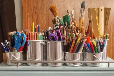 Image of 5 tin cans fulled with various art supplies, like scissors, paintbrushes, pencils, rulers, etc standing on a table.
