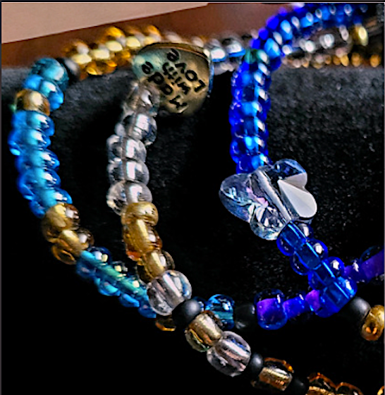 Image of 3 beaded bracelets, two blue, and one white with gold accents on each, resting on a black cylindrical showcase.