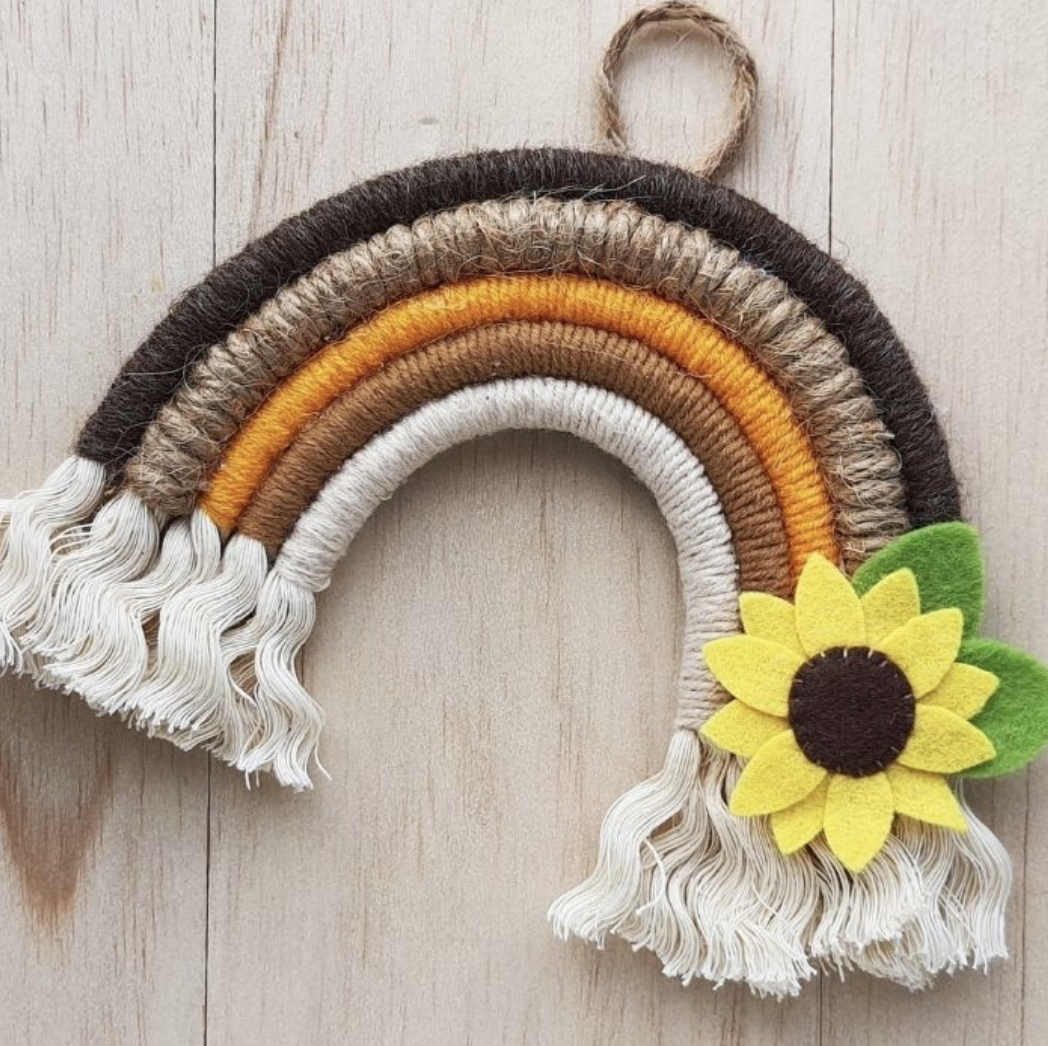 Image of a woven rainbow made of beige, brown, orange, light brown, and dark brown stripes with a felt sunflower on the bottom right end of it, wavy pieces of fabric flowing off the ends of the rainbow to resemble clouds.