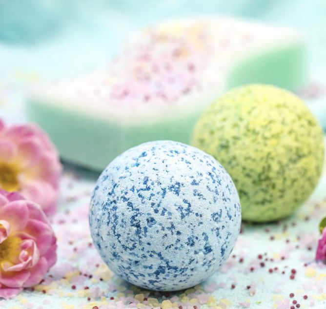 Image of a blue bath bomb and a green bath bomb resting on a table littered in seeds, salts, and flowers.