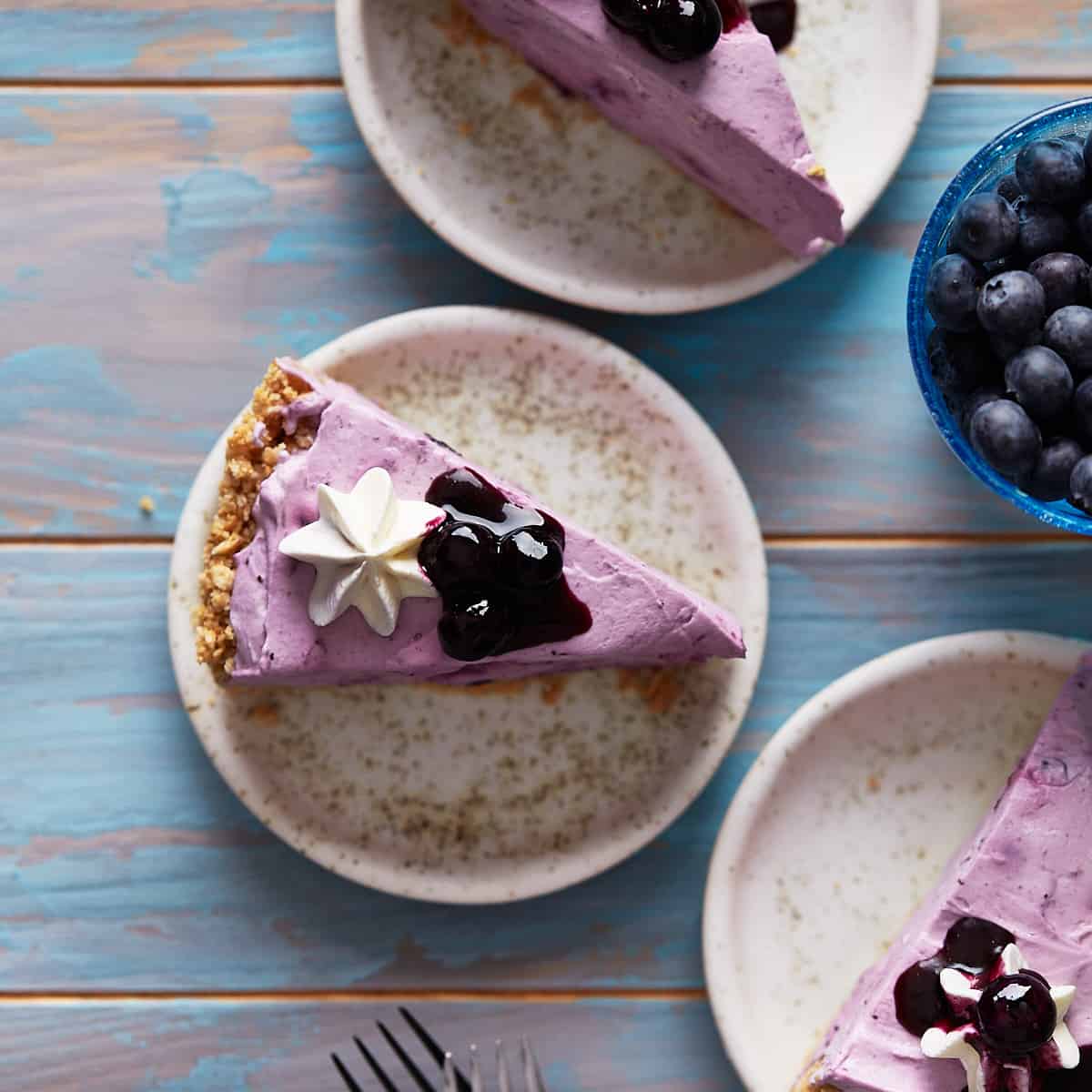 Image of blueberry cheesecakes on plates, with dollops of whipped cream atop each. There is a fork on the bottom and a bowl of blueberries on the right.