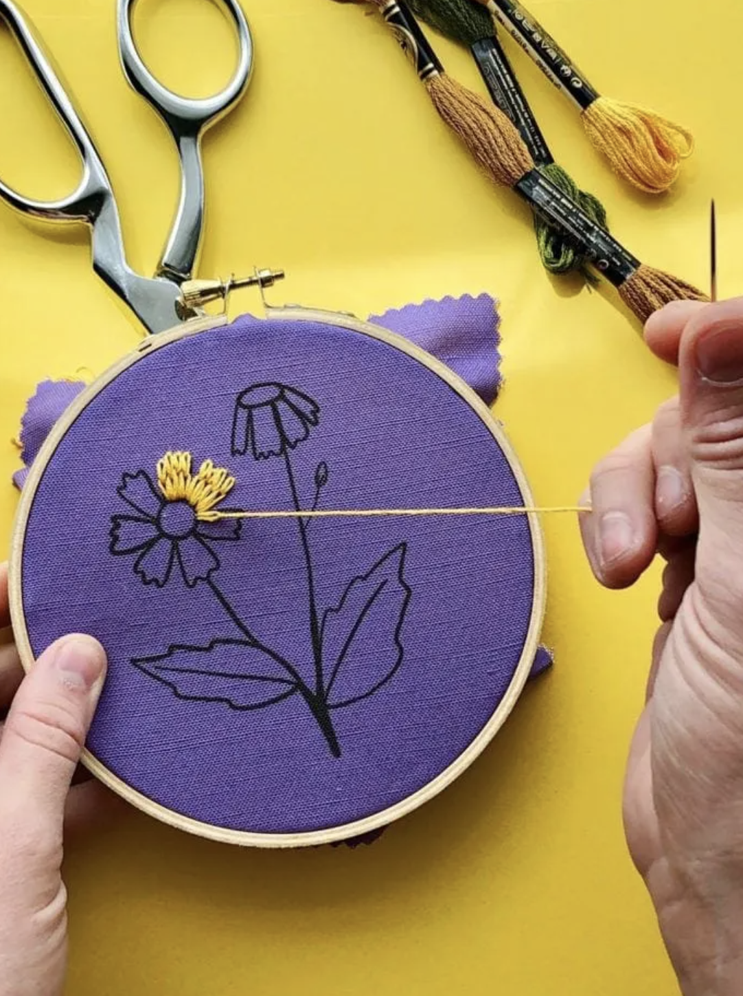 Two hands holding a purple embroidery circle, stitching a yellow flower onto it; only two petals are done. The background is yellow and has scissors and brushes laying behind the piece.