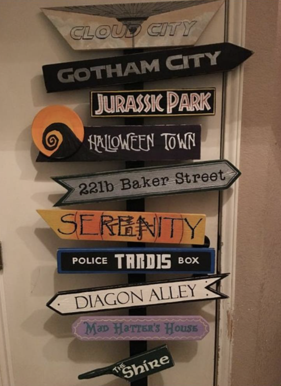 Image of a sign resting against the door with multiple arrows pointing to different locations, including, from top to bottom, cloud city, Gotham city, Jurassic Park, Halloween Town, 221b Baker Street, Serenity, Police Tardis Box, Diagon Alley, Mad Hatter's House, and The Shrine.