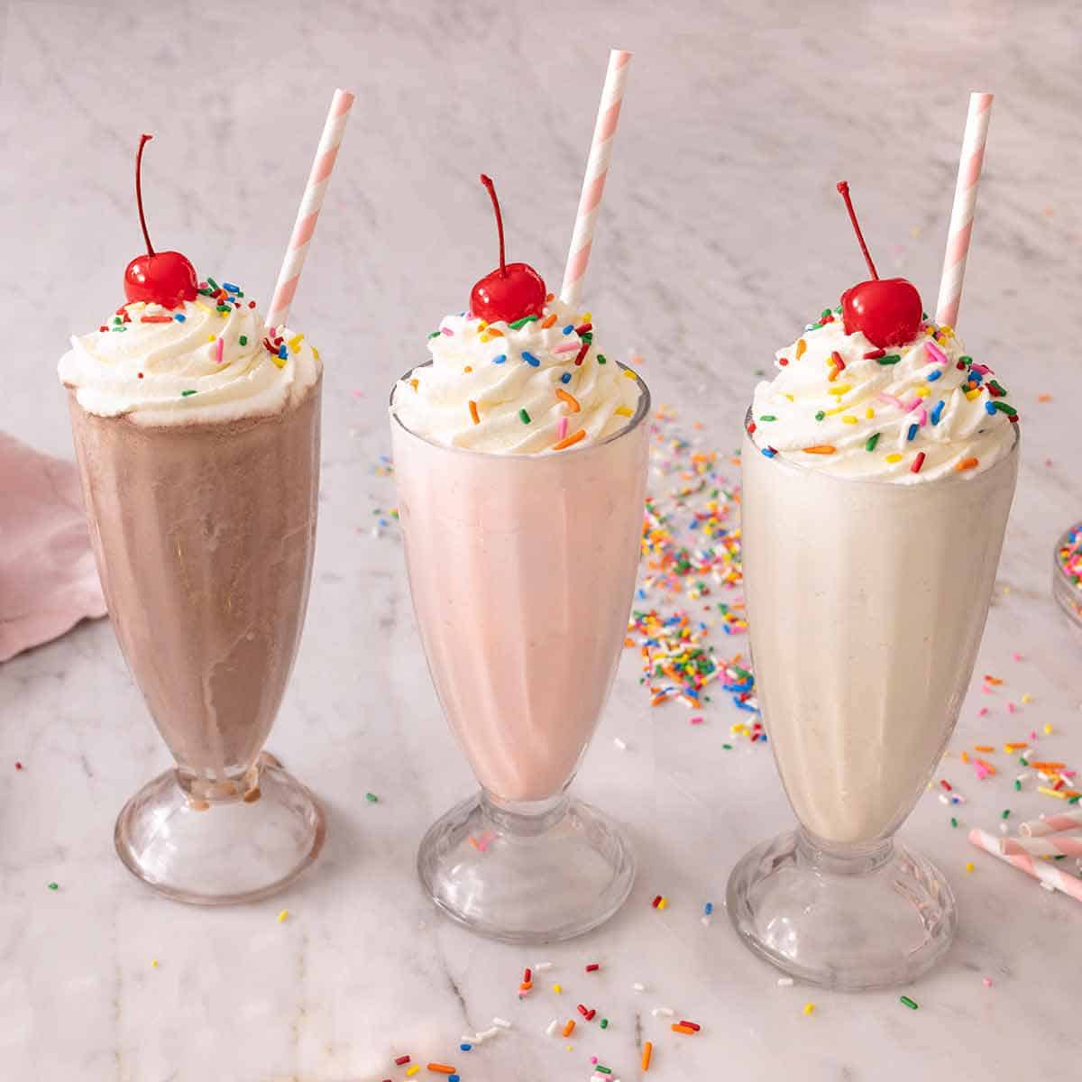 Image of a chocolate, strawberry, and vanilla milkshake with pink and white straws, whipped cream, rainbow sprinkles, and cherries on top, all in glasses atop a marble counter.
