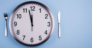 An analog clock with the time reading 11:57 AM, a fork placed at the left of the clock and a knife placed to the right of the clock. 