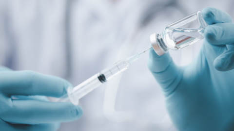 Photo of medical personnel with gloves using a syringe to get medicine out of a vial.