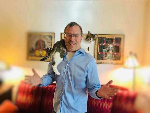 Image of Feline Behaviorist in a blue shirt with a white and gray cat climbing on his shoulders.