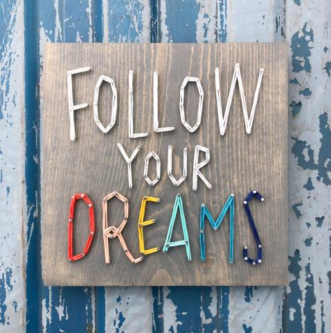Image of string art on a wood board reading "Follow Your Dreams," with the word "Dreams" in rainbow.