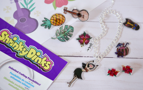 Image of a summer themed napkin with little transparent charm and pins on top of it, a logo reaing "Shrinky Dinks" off to the left over a purple paper laying atop the napkin and white wood background.