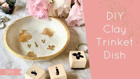 white and gold ceramic trinket dish on the left with flowers and small wooden keychains scattered beneath it, and a pink block on the right reads DIY Clay Trinket Dish.