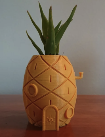 3D Printed Spongebob Pineapple House planter that is tinted yellow with what looks like thick green leaves sprouting from the top. It rests on a wood table.