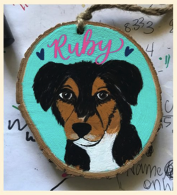 A wood slice with a black white and brown dog painted on it with a teal background and the name "Ruby" painted in script in pink above the dog's head.